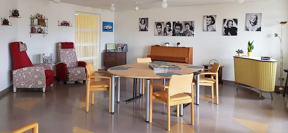 Runder Holztisch mit vier Stühlen in einem hellen Raum mit Sesseln, Kommode, Klavier und Schwarz-Weiß-Fotografien an der Wand.