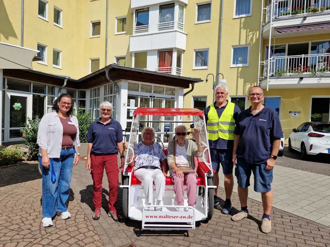 Sechs Personen stehen und sitzen vor einem gelben Wohngebäude um ein kleines weiß-rotes Fahrzeug mit der Aufschrift 'www.malteser-aw.de'