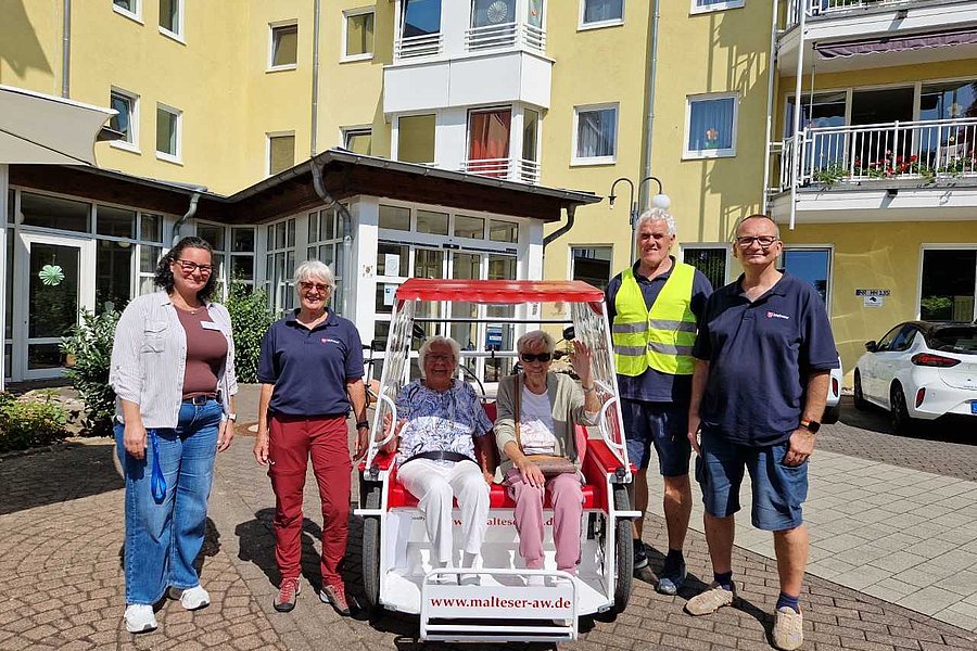 Sechs Personen stehen und sitzen vor einem gelben Wohngebäude um ein kleines weiß-rotes Fahrzeug mit der Aufschrift 'www.malteser-aw.de'