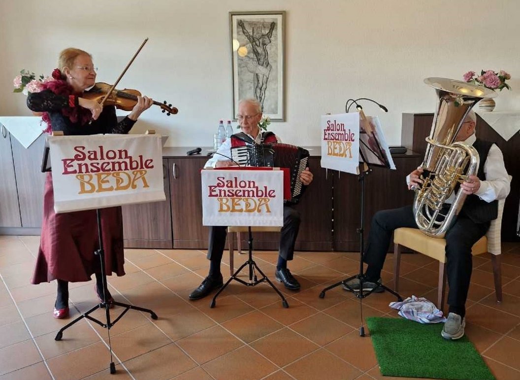 Drei Musiker des Salon Ensemble Beda spielen Geige, Akkordeon und Tuba in einem Raum mit Fliesenboden und einem Bild an der Wand.
