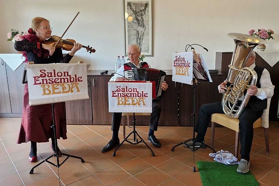 Drei Musiker des Salon Ensemble Beda spielen Geige, Akkordeon und Tuba in einem Raum mit Fliesenboden und einem Bild an der Wand.