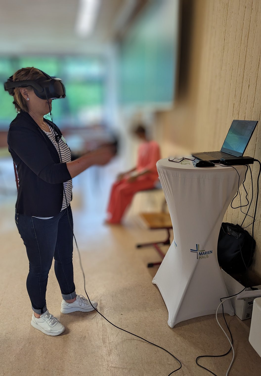 Frau mit VR-Brille und Controllern steht neben einem hohen Tisch mit Laptop, im Hintergrund sitzt eine weitere Person auf einer Bank.