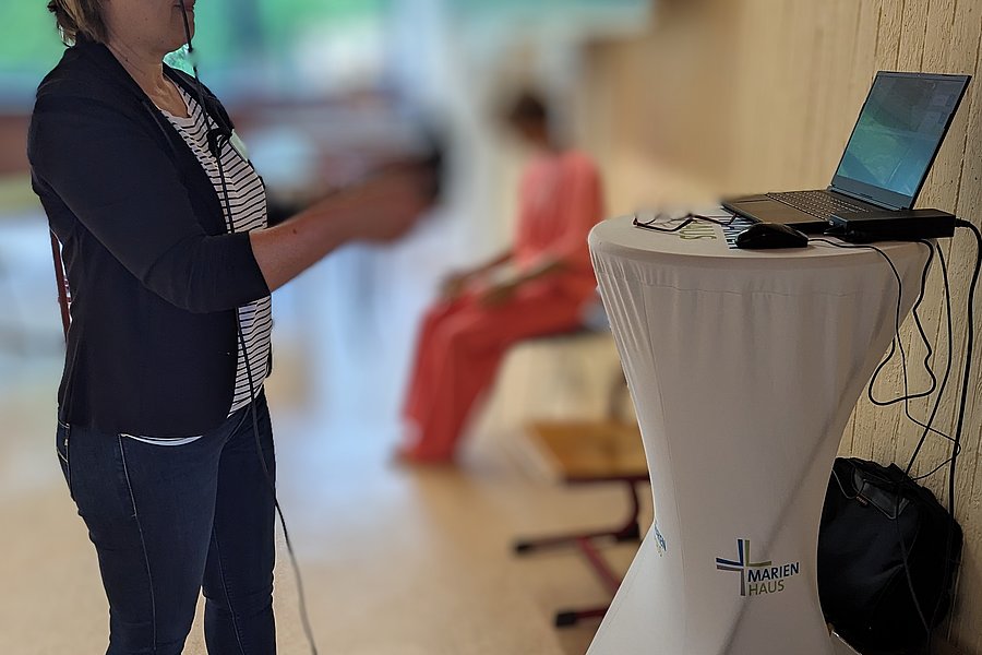 Frau mit VR-Brille und Controllern steht neben einem hohen Tisch mit Laptop, im Hintergrund sitzt eine weitere Person auf einer Bank.