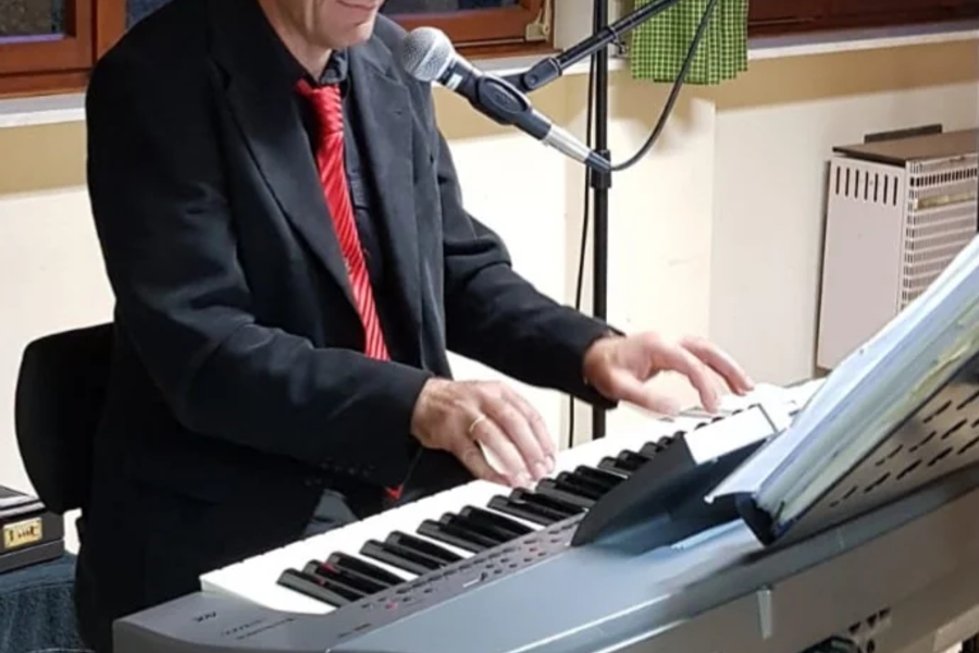 Mann in schwarzem Anzug und roter Krawatte spielt Keyboard der Marke Technics und singt in ein Mikrofon vor einem Fenster mit grünen Vorhängen.
