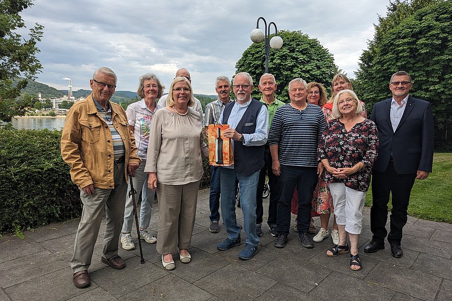 Zehn Personen stehen auf einem gepflasterten Weg vor Büschen und Bäumen, eine Person hält eine orange-schwarze Tasche.