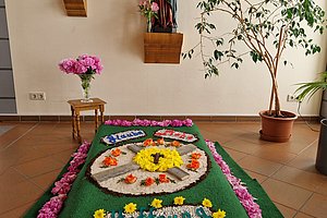 Grab mit grünem Grabteppich, verziert mit Blumenarrangements und umgeben von rosa Blumen, im Hintergrund zwei Heiligenfiguren und ein Bild an der Wand