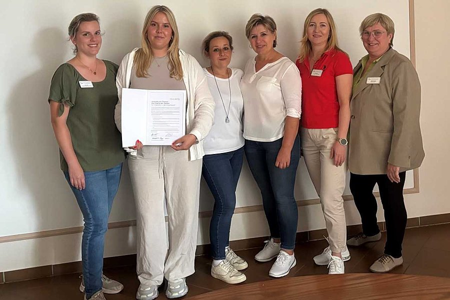 Sechs Frauen stehen nebeneinander vor einer Wand, eine hält ein Dokument mit Text, alle tragen legere Kleidung und Namensschilder.