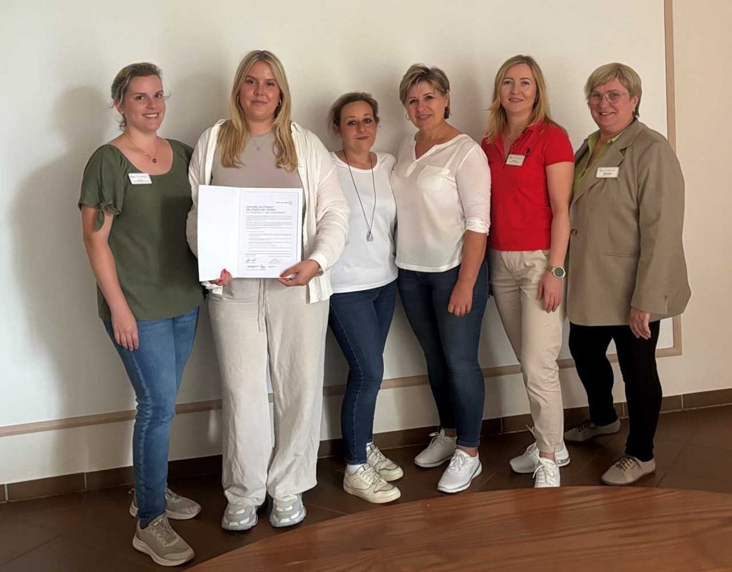 Sechs Frauen stehen nebeneinander vor einer Wand, eine hält ein Dokument mit Text, alle tragen legere Kleidung und Namensschilder.