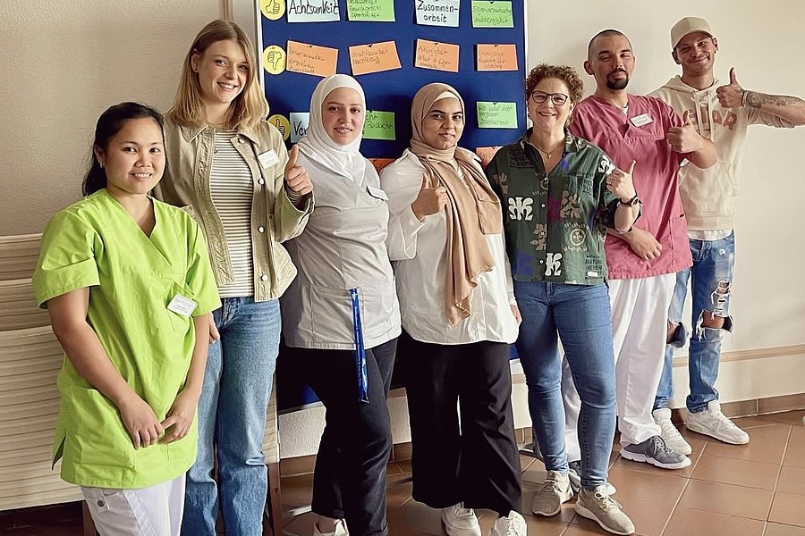 Sieben Personen stehen vor einer Wand mit einem blauen Pinnbrett, auf dem bunte Zettel befestigt sind. Über der Wand steht 'BSS - COACHING & TRAINING'.