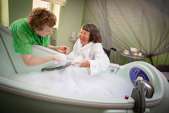 Person in weißem Bademantel sitzt in einer Badewanne mit Schaum, eine andere Person in grünem Oberteil beugt sich über die Wanne und hält die Hand der sitzenden Person.