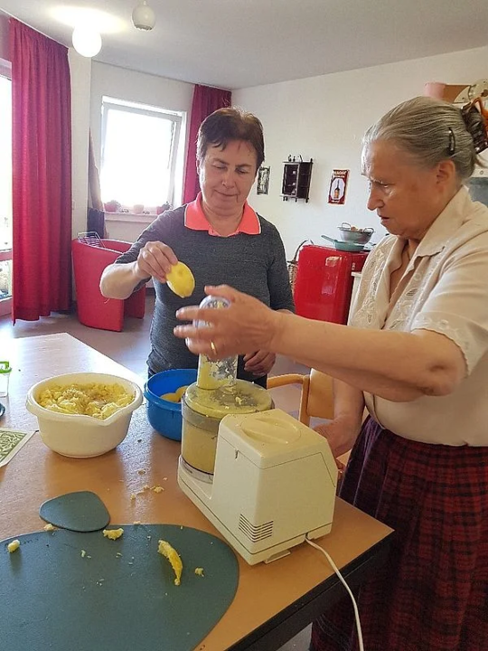 Zwei Personen bereiten in einer Küche mit rotem Kühlschrank und roten Vorhängen Essen zu, eine bedient eine Küchenmaschine, die andere hält eine Kartoffel.