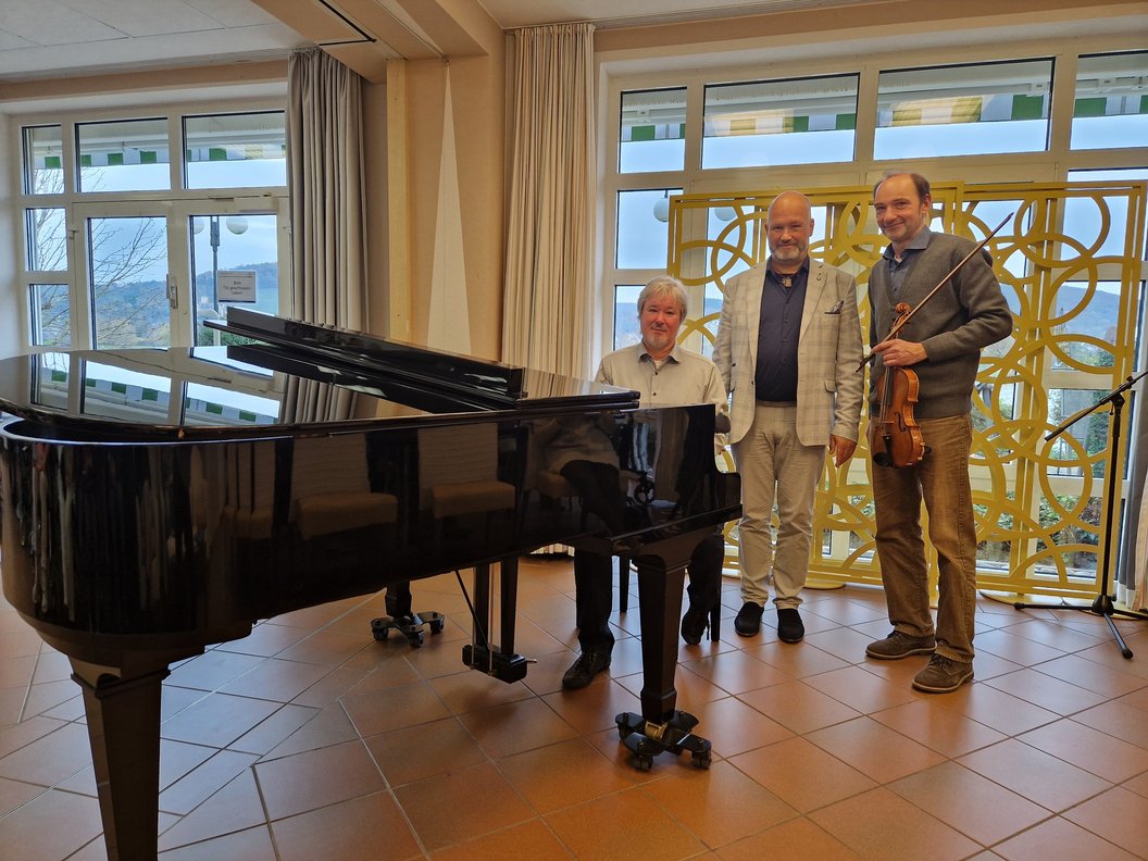 Großer schwarzer Flügelklavier im Vordergrund, drei Personen stehen dahinter, eine hält eine Geige, helle Gardinen und große Fenster im Hintergrund.