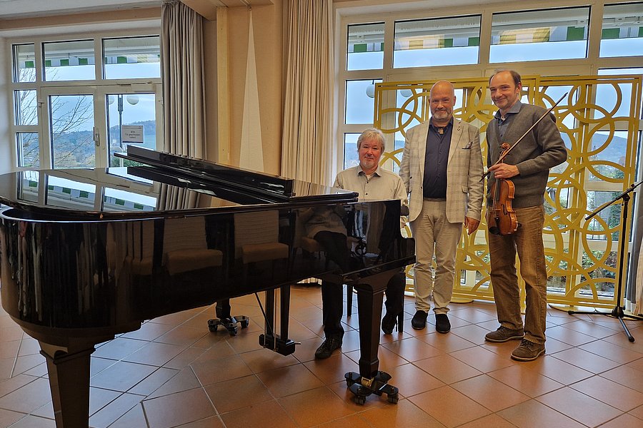 Großer schwarzer Flügelklavier im Vordergrund, drei Personen stehen dahinter, eine hält eine Geige, helle Gardinen und große Fenster im Hintergrund.