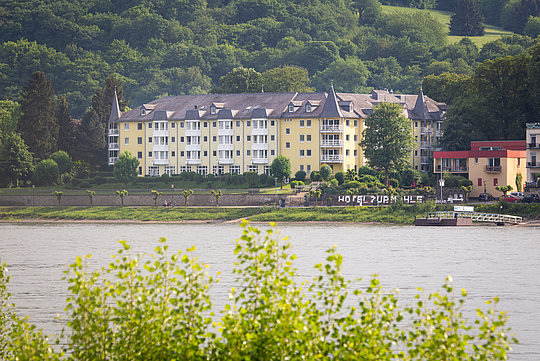 Großes gelbes Hotelgebäude mit mehreren Etagen und Türmen am Ufer eines Flusses, umgeben von grünen Bäumen und Hügeln.