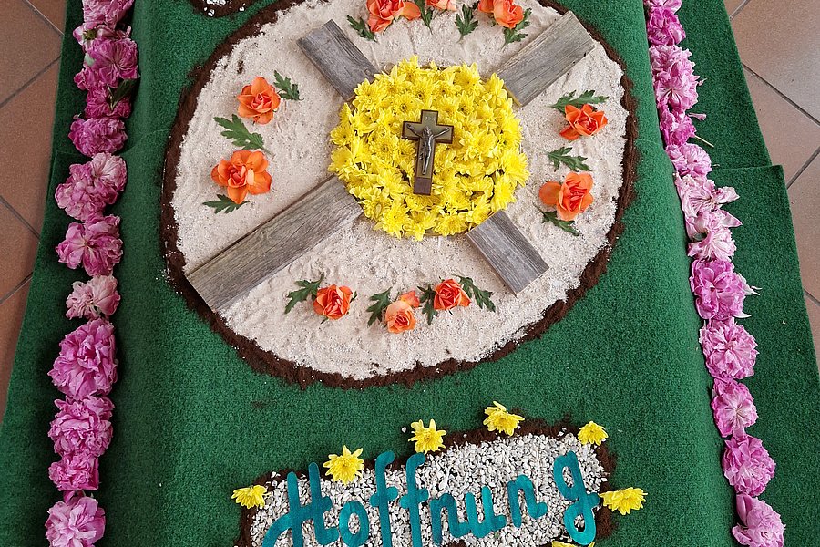 Grüner rechteckiger Teppich mit Blumenarrangements in der Mitte und an den Rändern, mittig ein gelber Kreis mit einem Kreuz, umgeben von weißen und orangen Blüten, oben zwei ovale Blumenfelder mit den Worten 'Glaube' und 'Liebe', unten ein Blumenfeld mit dem Wort 'Hoffnung'