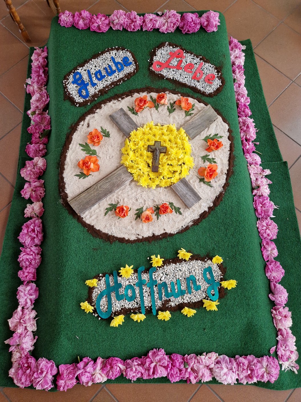 Grüner rechteckiger Teppich mit Blumenarrangements in der Mitte und an den Rändern, mittig ein gelber Kreis mit einem Kreuz, umgeben von weißen und orangen Blüten, oben zwei ovale Blumenfelder mit den Worten 'Glaube' und 'Liebe', unten ein Blumenfeld mit dem Wort 'Hoffnung'