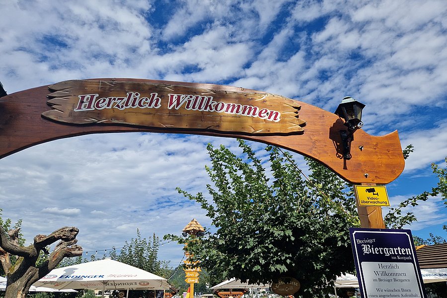 Holzbogen mit dem Schriftzug 'Herzlich Willkommen' über einem Biergarten mit Bäumen, Sonnenschirmen und blauem Himmel mit Wolken.