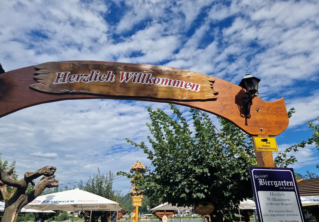 Holzbogen mit dem Schriftzug 'Herzlich Willkommen' über einem Biergarten mit Bäumen, Sonnenschirmen und blauem Himmel mit Wolken.