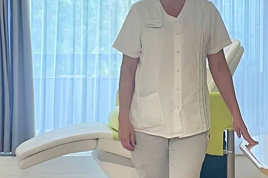 Eine Person in weißer medizinischer Kleidung steht in einem hellen Raum mit großen Fenstern und einer Liege im Hintergrund.