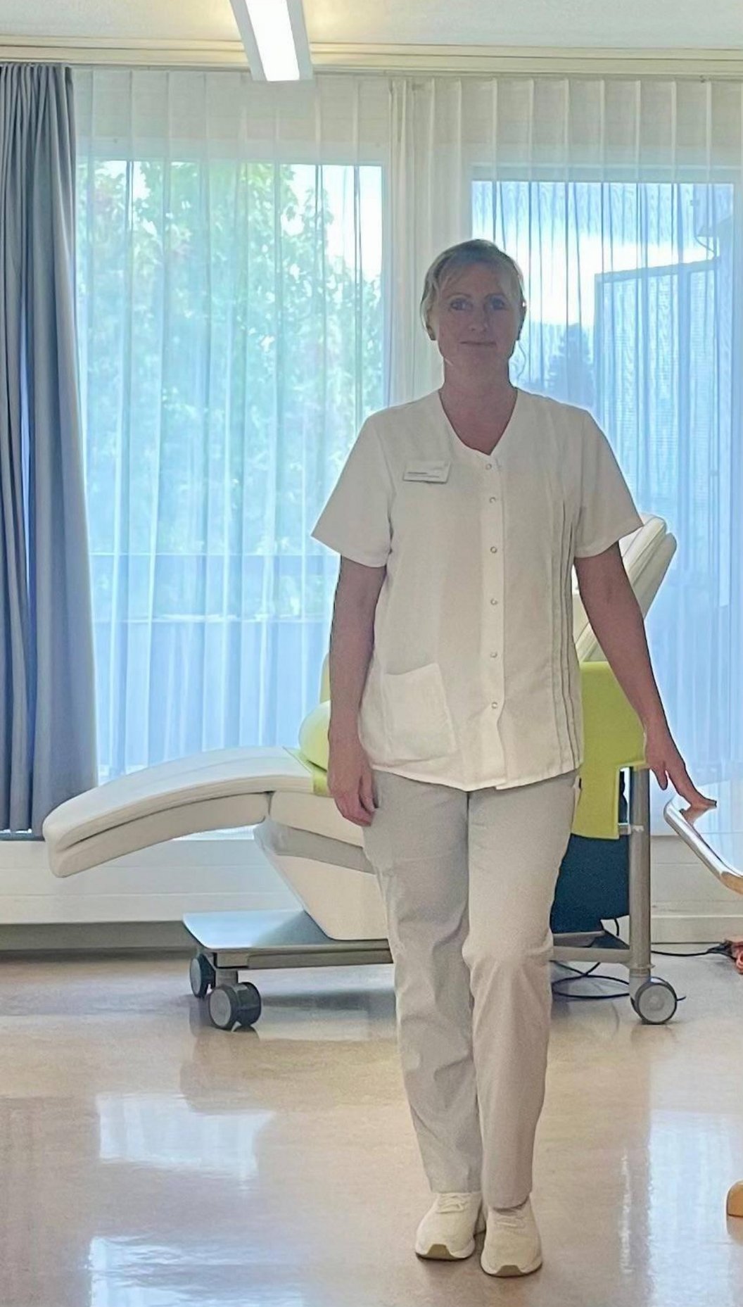 Eine Person in weißer medizinischer Kleidung steht in einem hellen Raum mit großen Fenstern und einer Liege im Hintergrund.