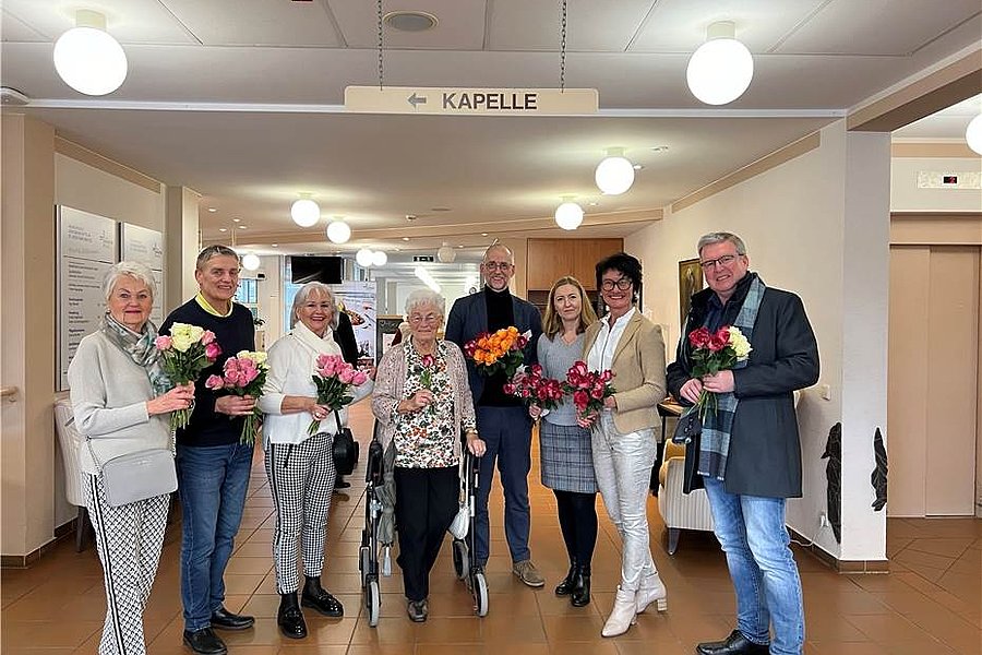 Acht Personen stehen in einem Flur unter einem Schild mit der Aufschrift 'Kapelle', jede hält einen Strauß bunter Blumen.
