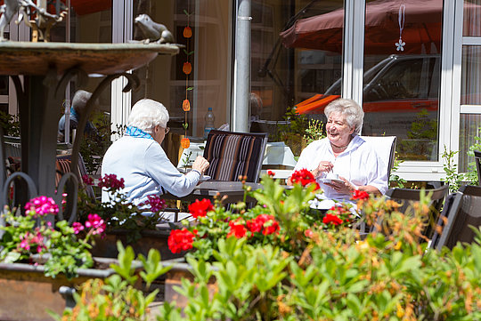 Zwei ältere Frauen sitzen an einem Tisch im Freien, umgeben von bunten Blumen und Pflanzen, vor einem Gebäude mit großen Fenstern.