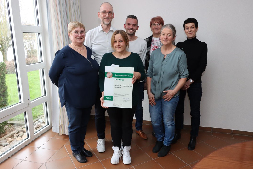 Sieben Personen stehen in einem Raum mit Fliesenboden und weißen Wänden, eine Person hält ein Zertifikat mit grünem Logo und Text.