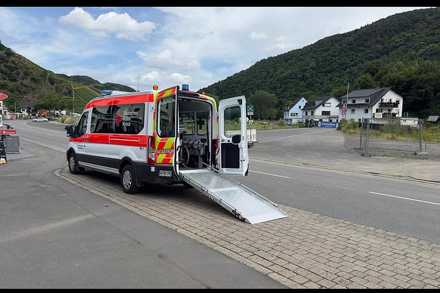Rettungswagen mit geöffneter Heckklappe und ausgefahrener Rampe auf einem Parkplatz, umgeben von Bergen und Häusern.