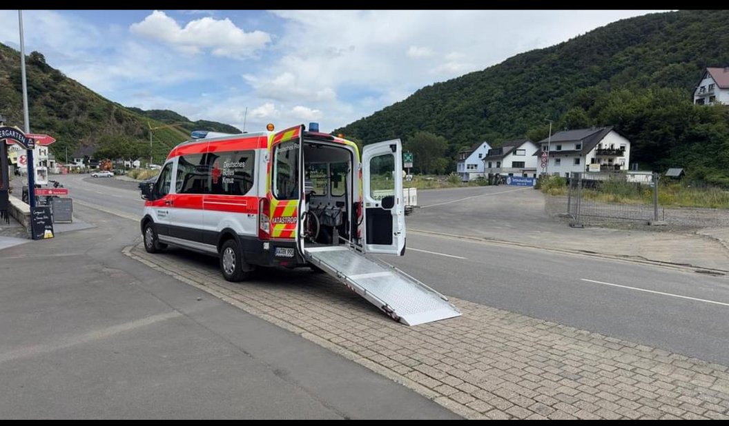 Rettungswagen mit geöffneter Heckklappe und ausgefahrener Rampe auf einem Parkplatz, umgeben von Bergen und Häusern.