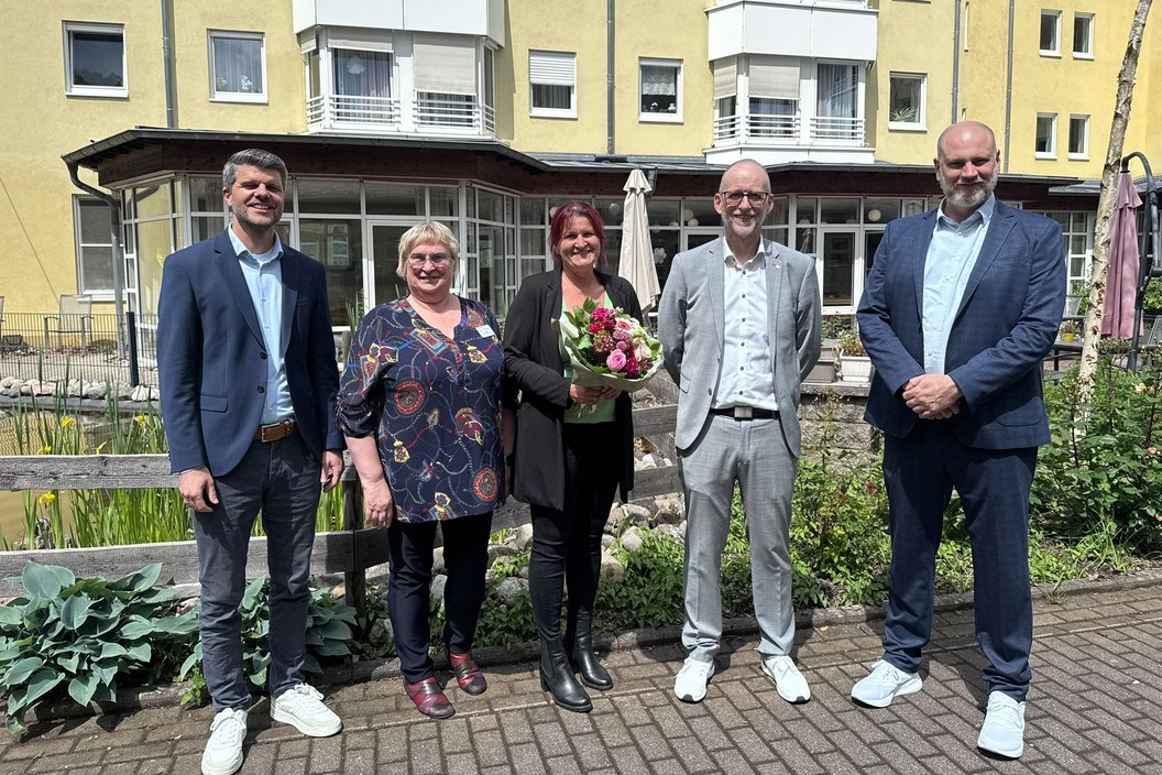 Fünf Personen posieren im Freien, umgeben von Pflanzen und einem Gebäude, einige halten Blumen.