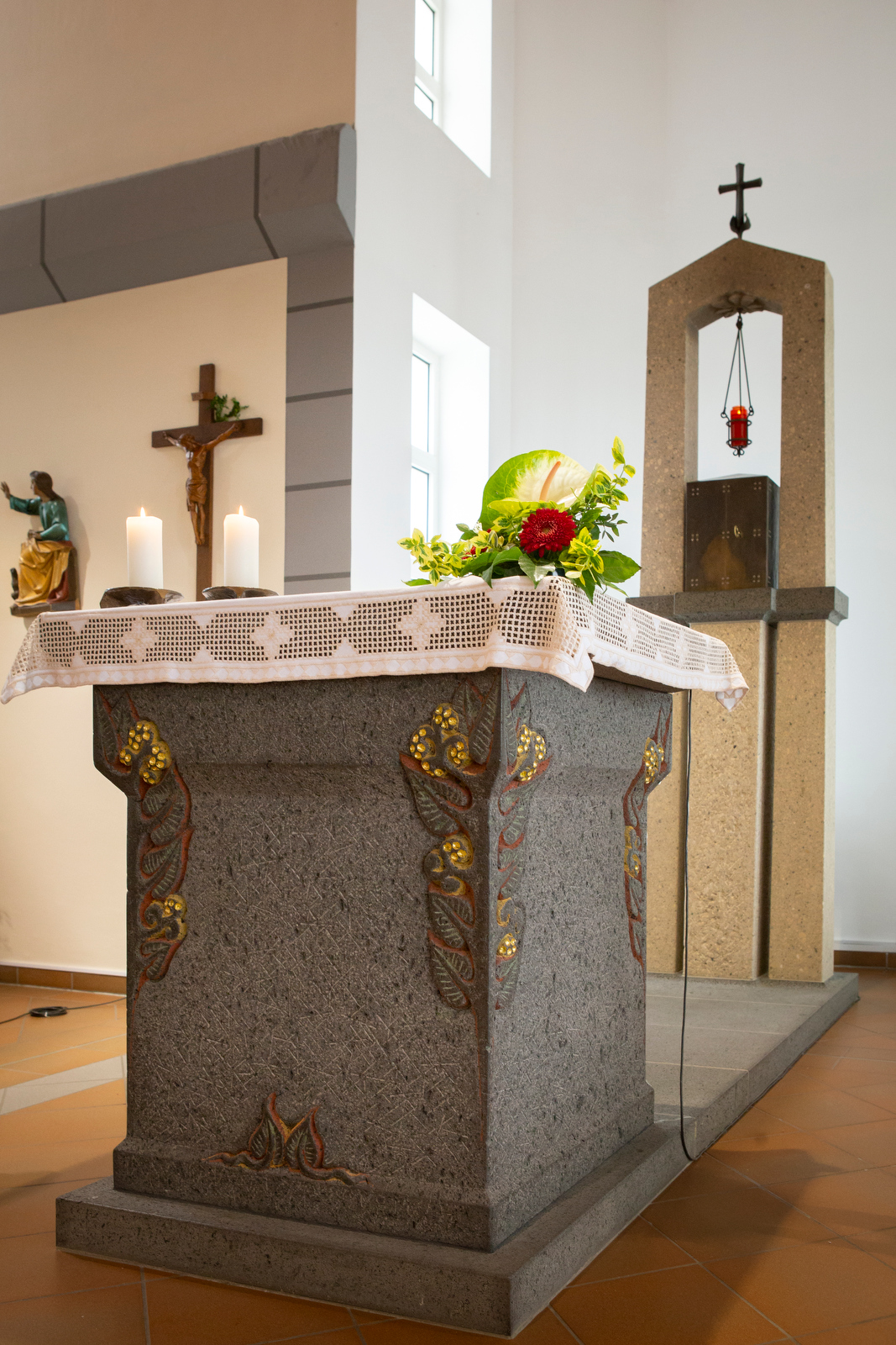 Steinerner Altar mit weißer Spitze, Blumen und Kerzen in einer Kirche, im Hintergrund ein Kreuz und eine Statue.