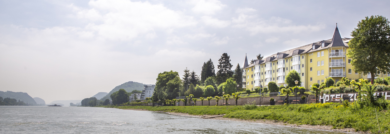 Gelbes Hotelgebäude mit spitzem Turm neben einem Fluss, bewölkter Himmel und bewaldete Hügel im Hintergrund.
