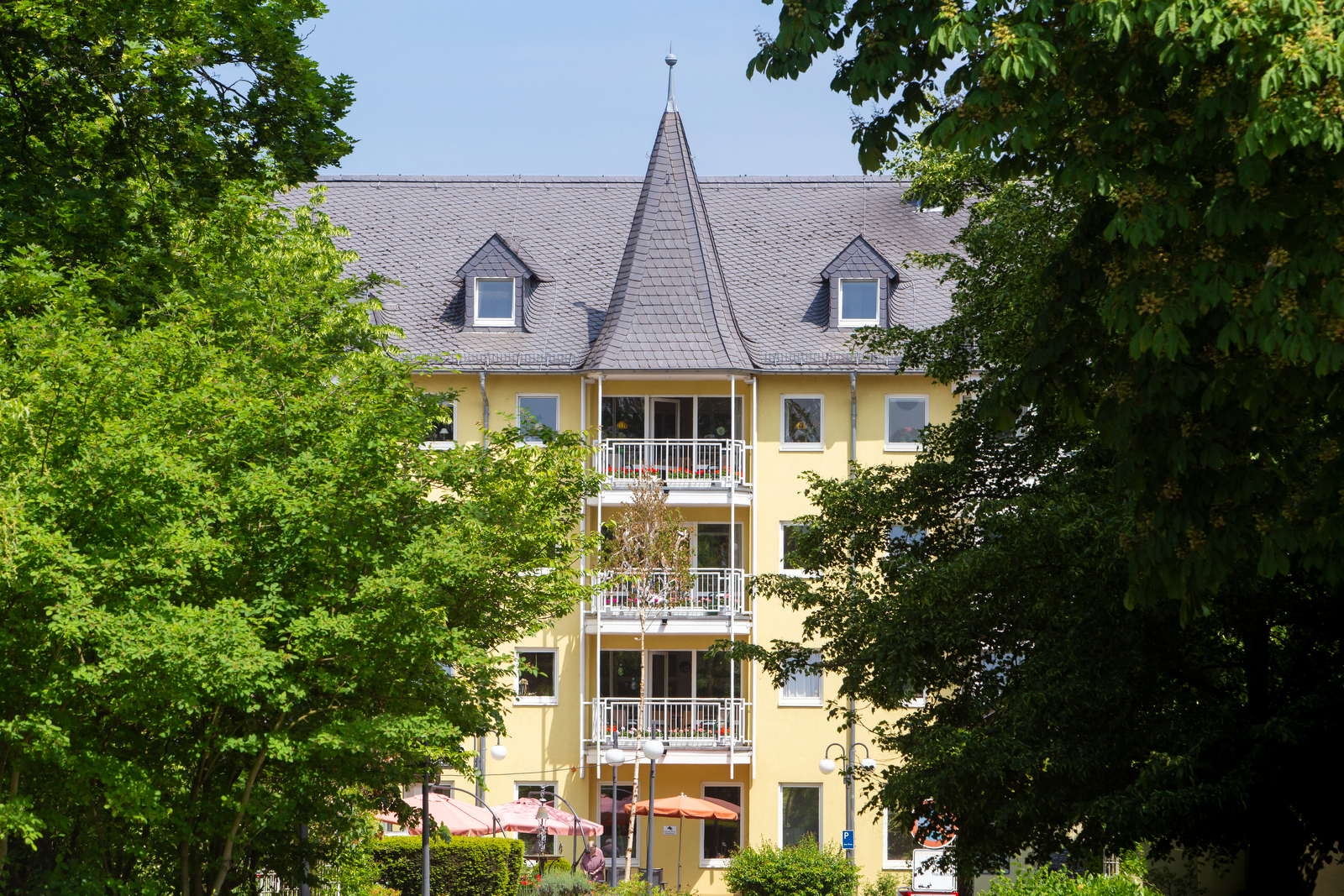 Gelbes mehrstöckiges Gebäude mit spitzem Dach und Balkonen, umgeben von grünen Bäumen unter blauem Himmel.