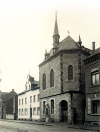 Straße mit mehreren Gebäuden, darunter ein steinernes Kirchengebäude mit spitzem Turm und gotischen Fenstern.