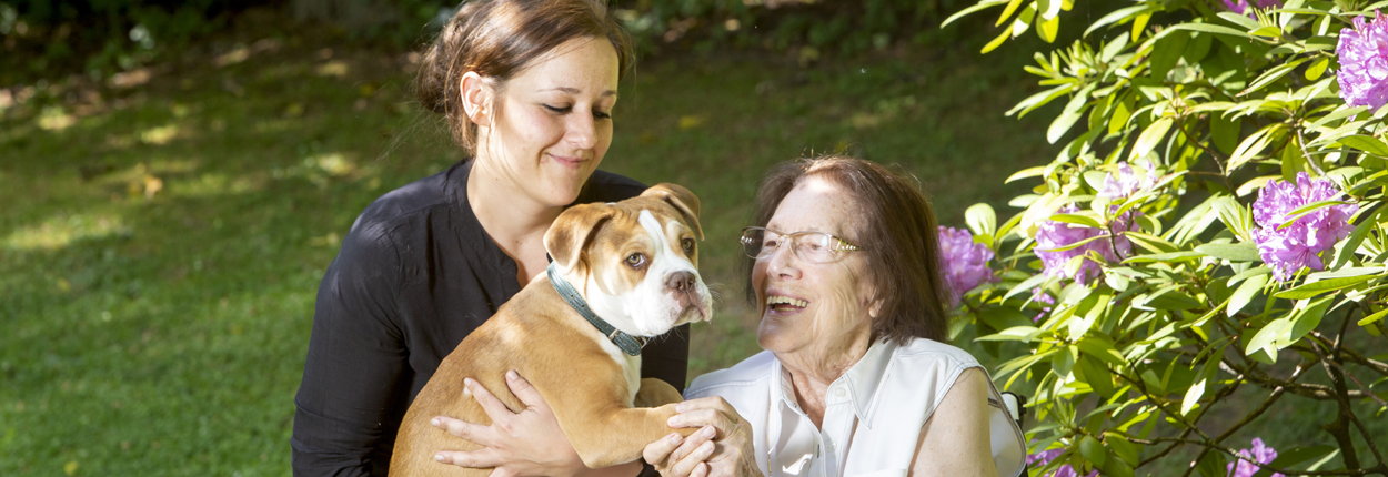 Zwei Personen sitzen im Garten, eine hält einen braunen Hund im Arm, umgeben von grünen Pflanzen und lila Blüten.