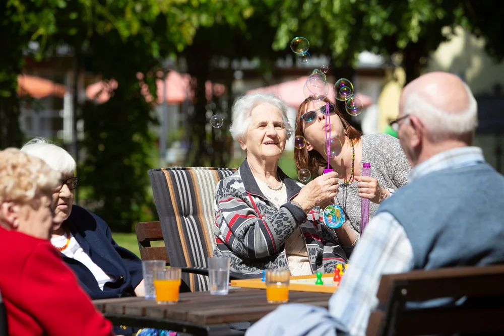 Mehrere ältere Menschen sitzen an einem Holztisch im Freien, auf dem Gläser mit orangefarbigen Getränken und ein Spielbrett stehen.