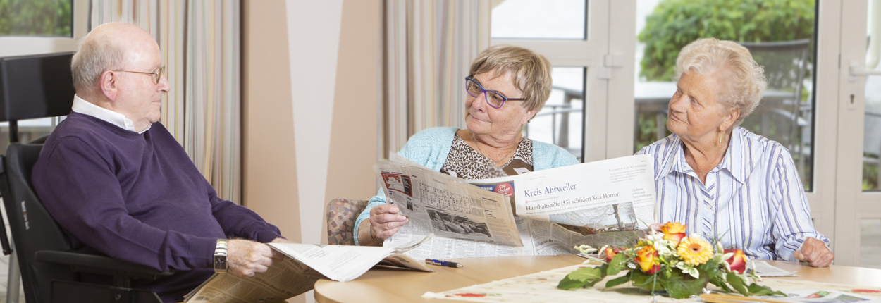 Drei ältere Personen sitzen an einem Tisch mit Blumenarrangement, zwei lesen Zeitungen, eine trägt einen dunkelblauen Pullover.