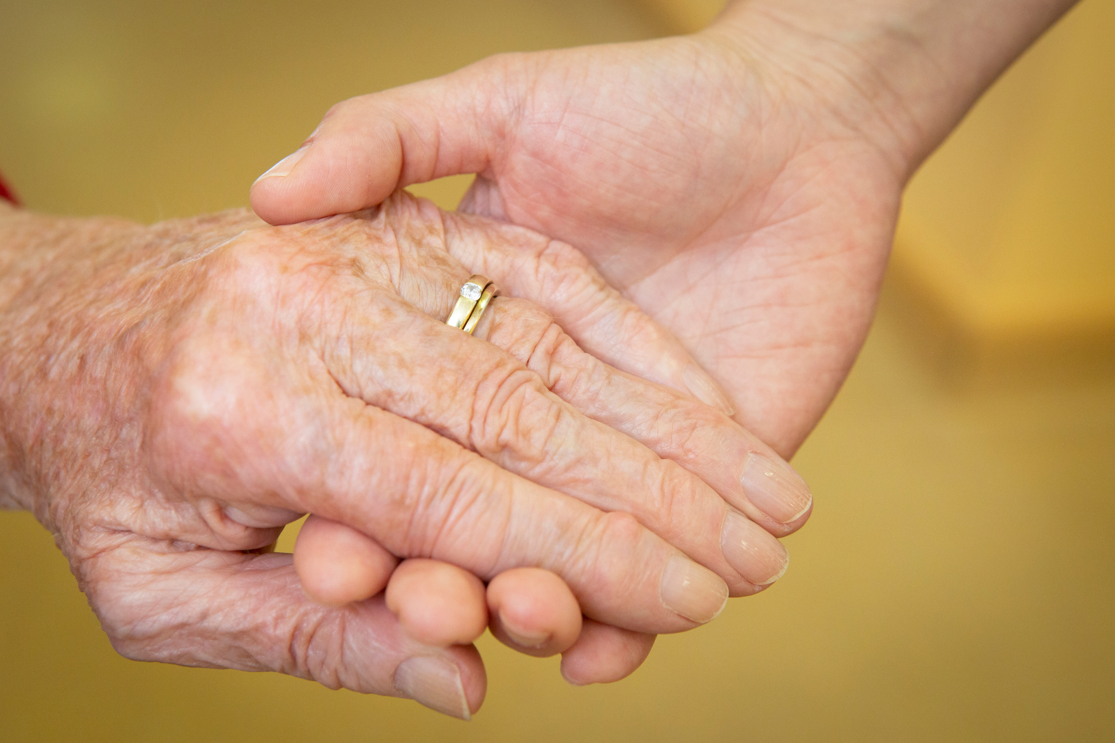 Junge Hand hält ältere Hand mit Ehering, beide Hände sind sichtbar, Hintergrund unscharf.
