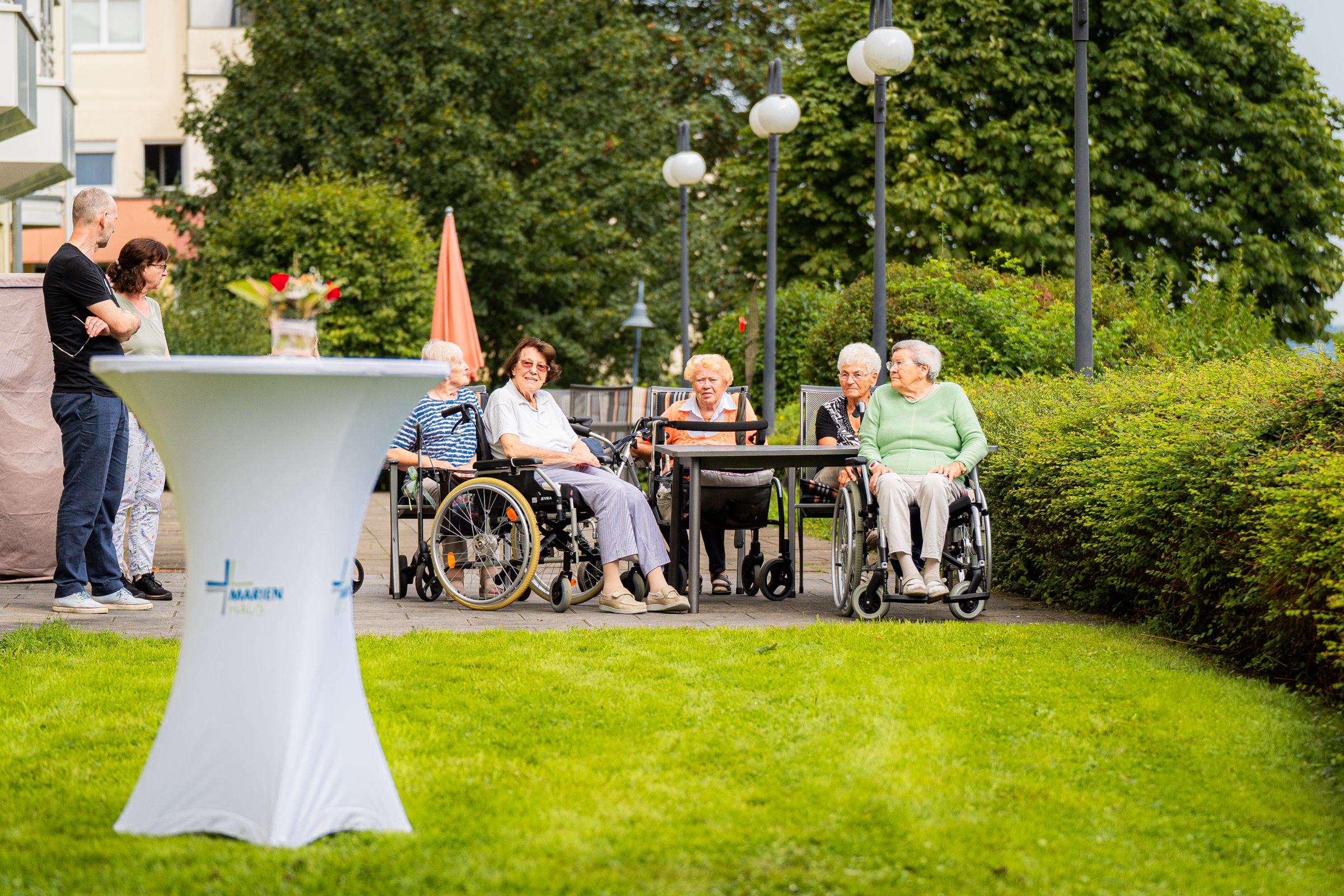 Mehrere ältere Menschen, einige im Rollstuhl, sitzen an einem Tisch im Garten. Zwei Personen stehen neben einem hohen Stehtisch mit einer Vase und roten Blumen.