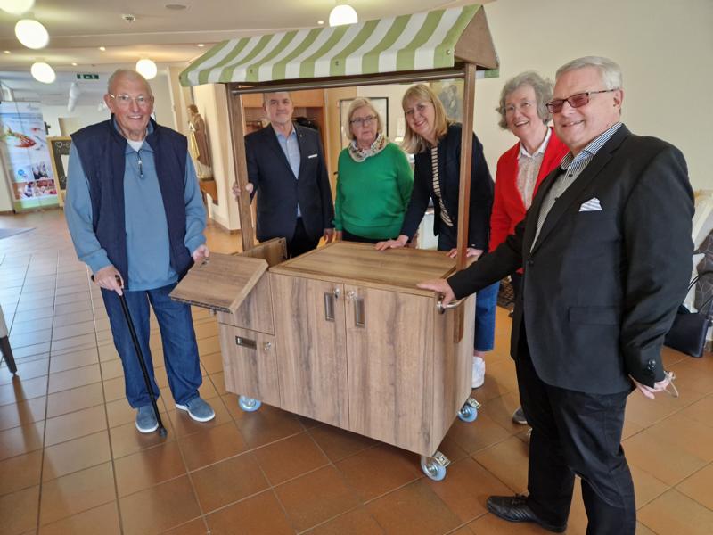 Sechs Personen stehen um einen fahrbaren Holztisch mit einem kleinen Dach, der an einen Marktstand erinnert, in einem Innenraum mit Fliesenboden.