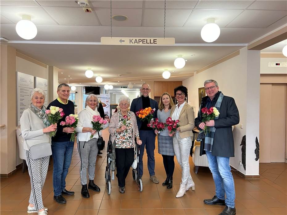 Acht Personen stehen in einem Flur unter einem Schild mit der Aufschrift 'Kapelle', jede hält einen Strauß bunter Blumen.