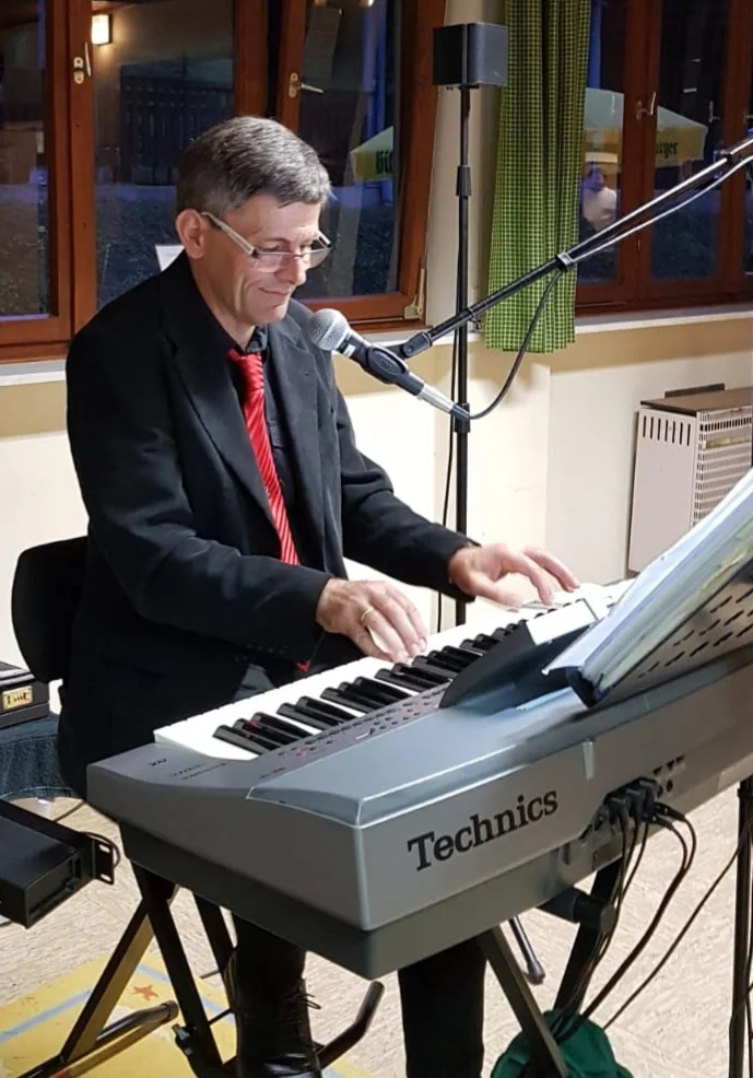 Mann in schwarzem Anzug und roter Krawatte spielt Keyboard der Marke Technics und singt in ein Mikrofon vor einem Fenster mit grünen Vorhängen.